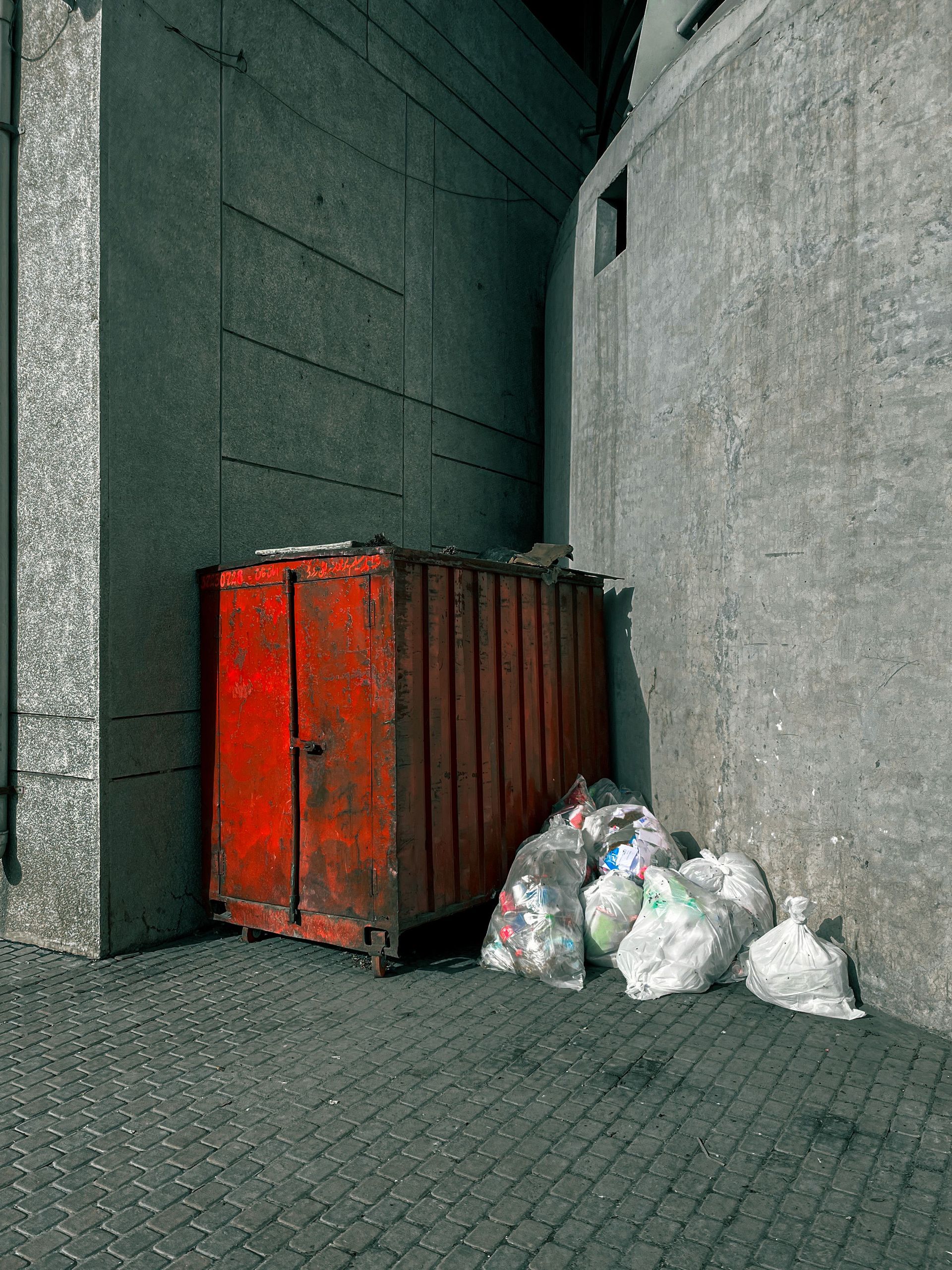 A rusty red dumpster with piled trash bags on an urban sidewalk representing waste management.