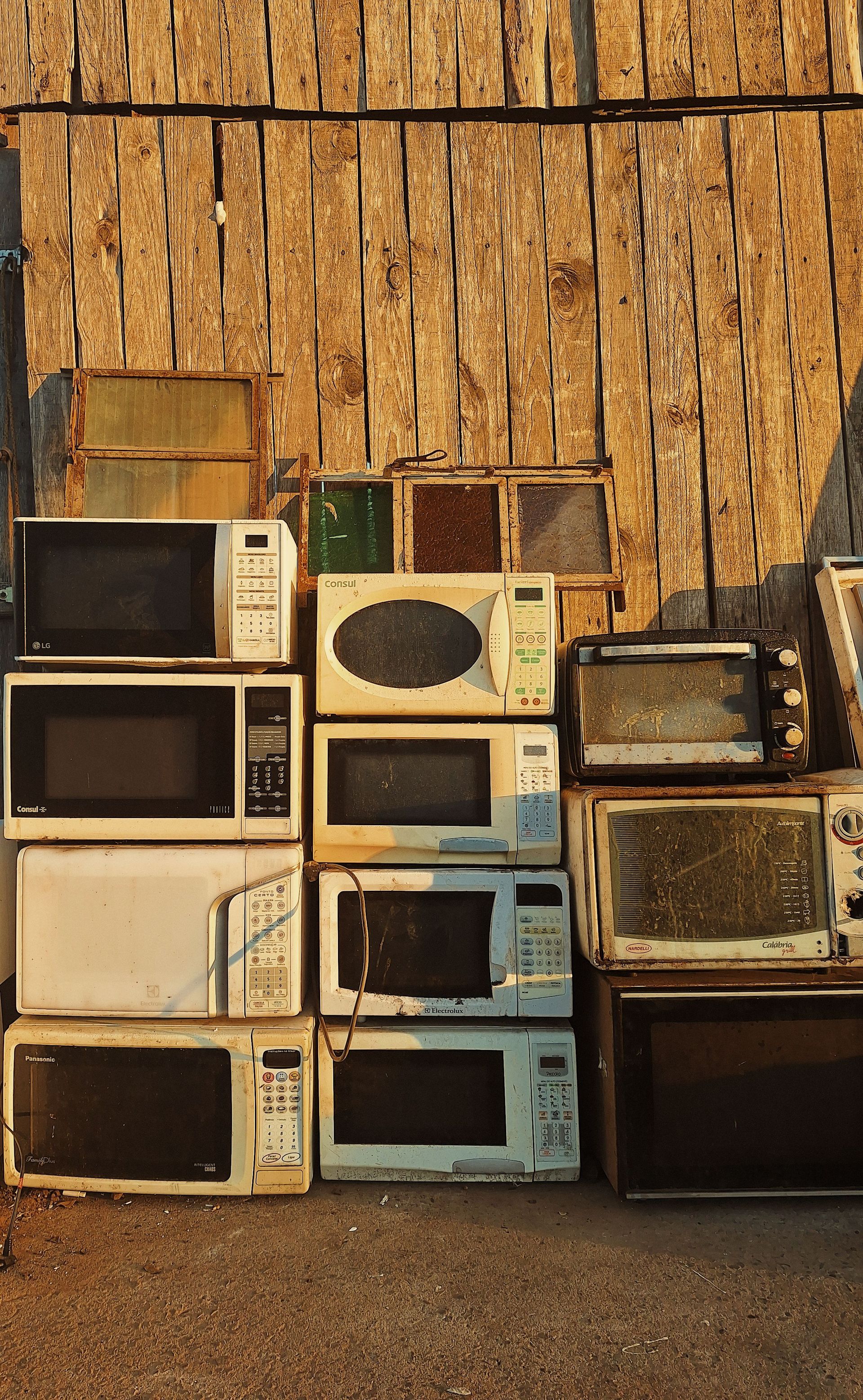 A stack of old microwaves placed outdoors against a rustic wooden wall in warm sunlight.